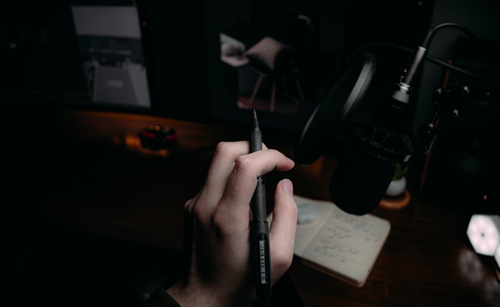 A hand holding a pen over an open notebook at a dimly lit desk.